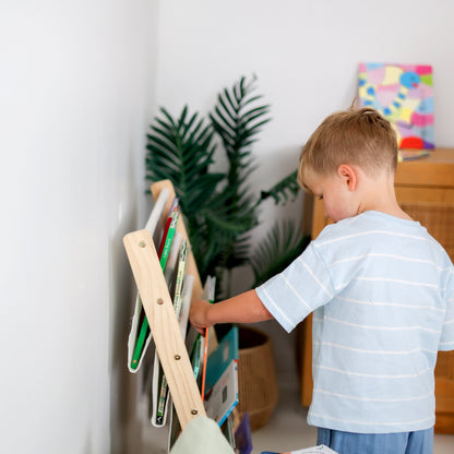 Child interacting with a wooden bookshelf with Spanish books in a room with plants and a colorful painting.